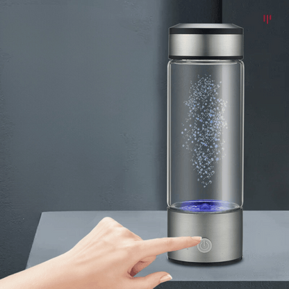 Glass water bottle with a silver base and black lid on a dark surface, with a hand touching the base.